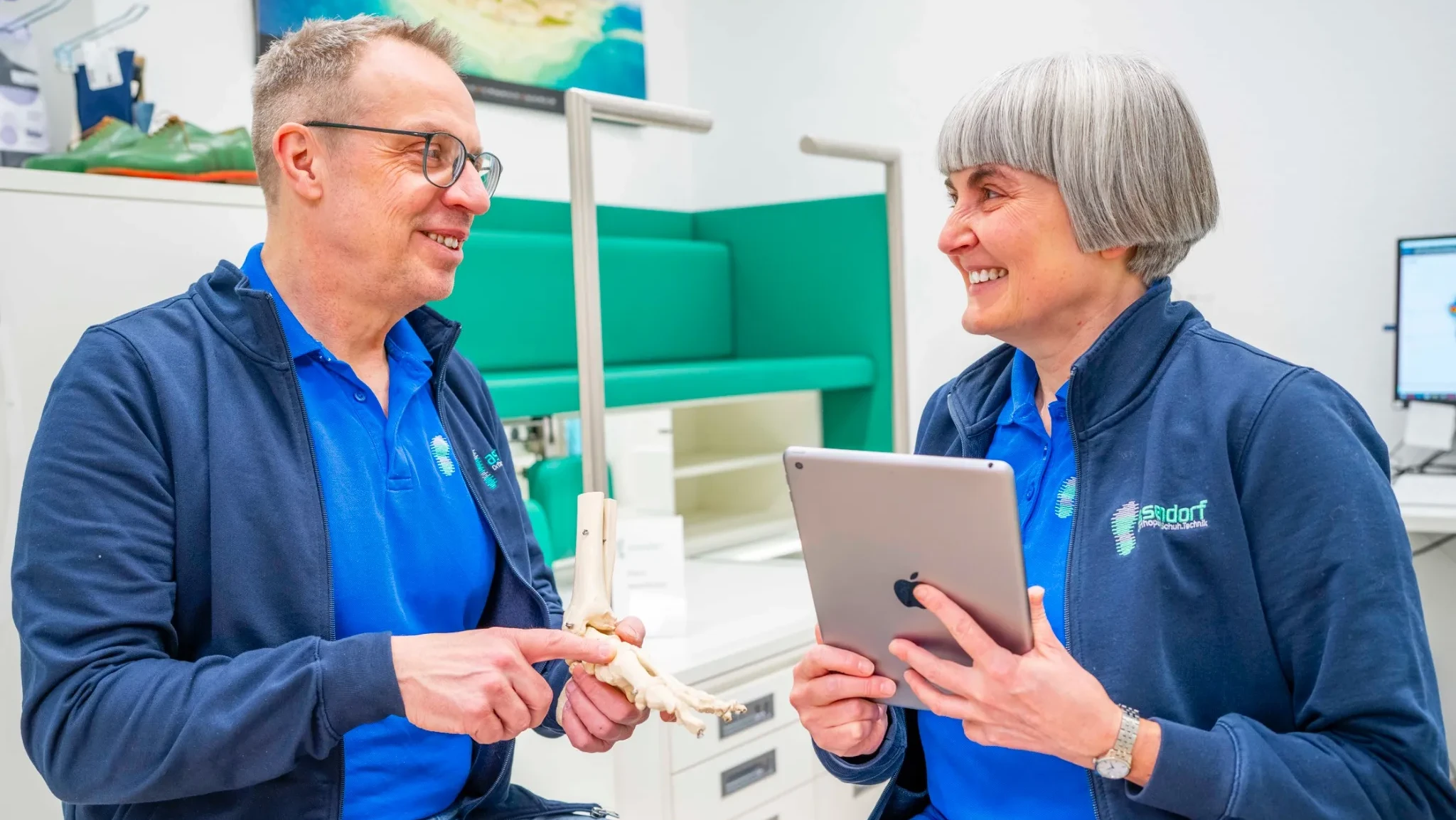 Jens und Karin Asendorf in blauer Asendorf-Kleidung sitzen in einem sportmedizinischen Büro; einer hält ein Fußknochenmodell, die andere ein Tablet. Beide lächeln und sind in ein Gespräch vertieft.