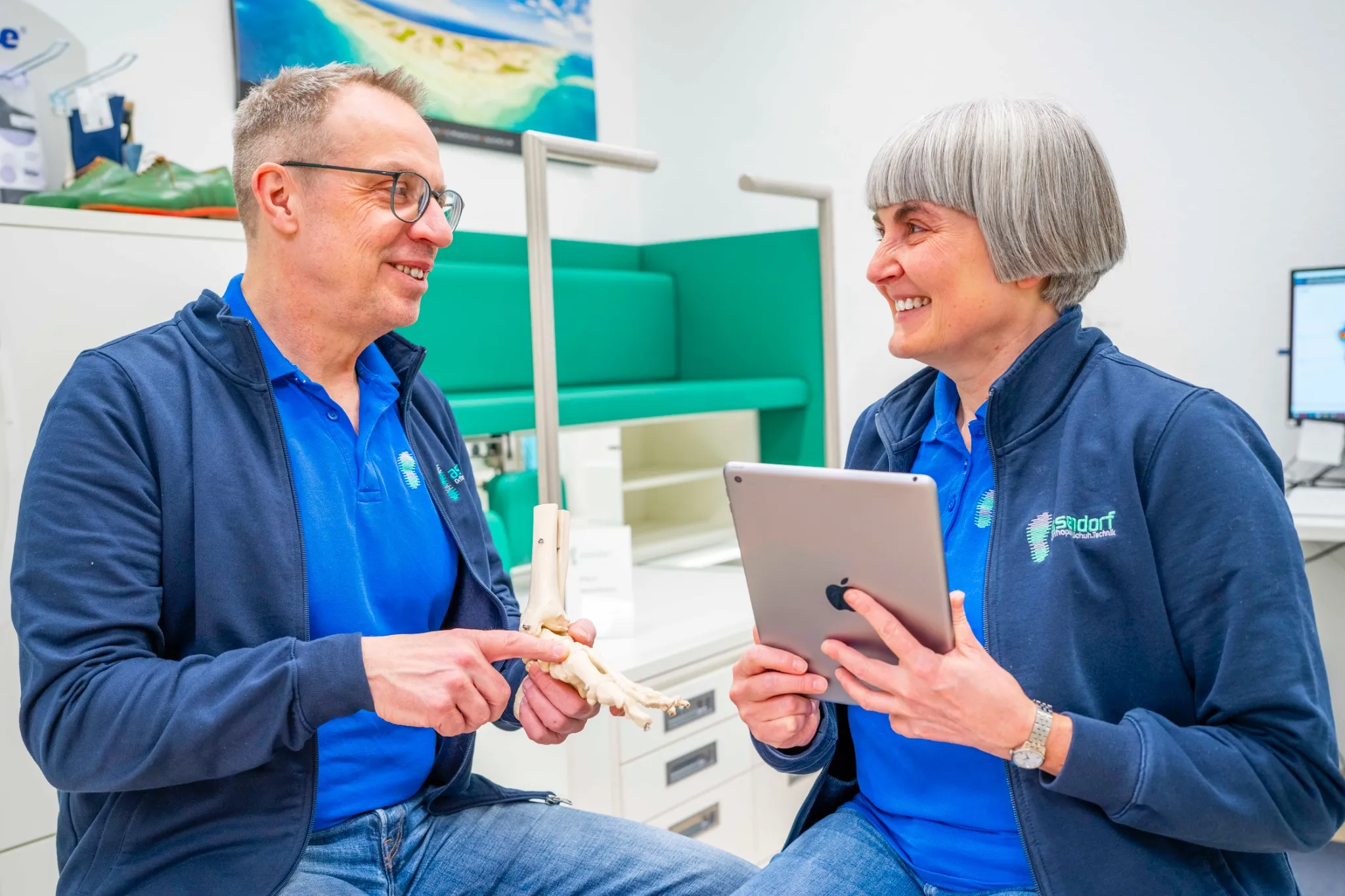 asendorf-orthopaedie-schuh-technik-bremen-gespraech Jens und Karin Asendorf in blauer Asendorf-Kleidung sitzen in einem sportmedizinischen Büro; einer hält ein Fußknochenmodell, die andere ein Tablet. Beide lächeln und sind in ein Gespräch vertieft.
