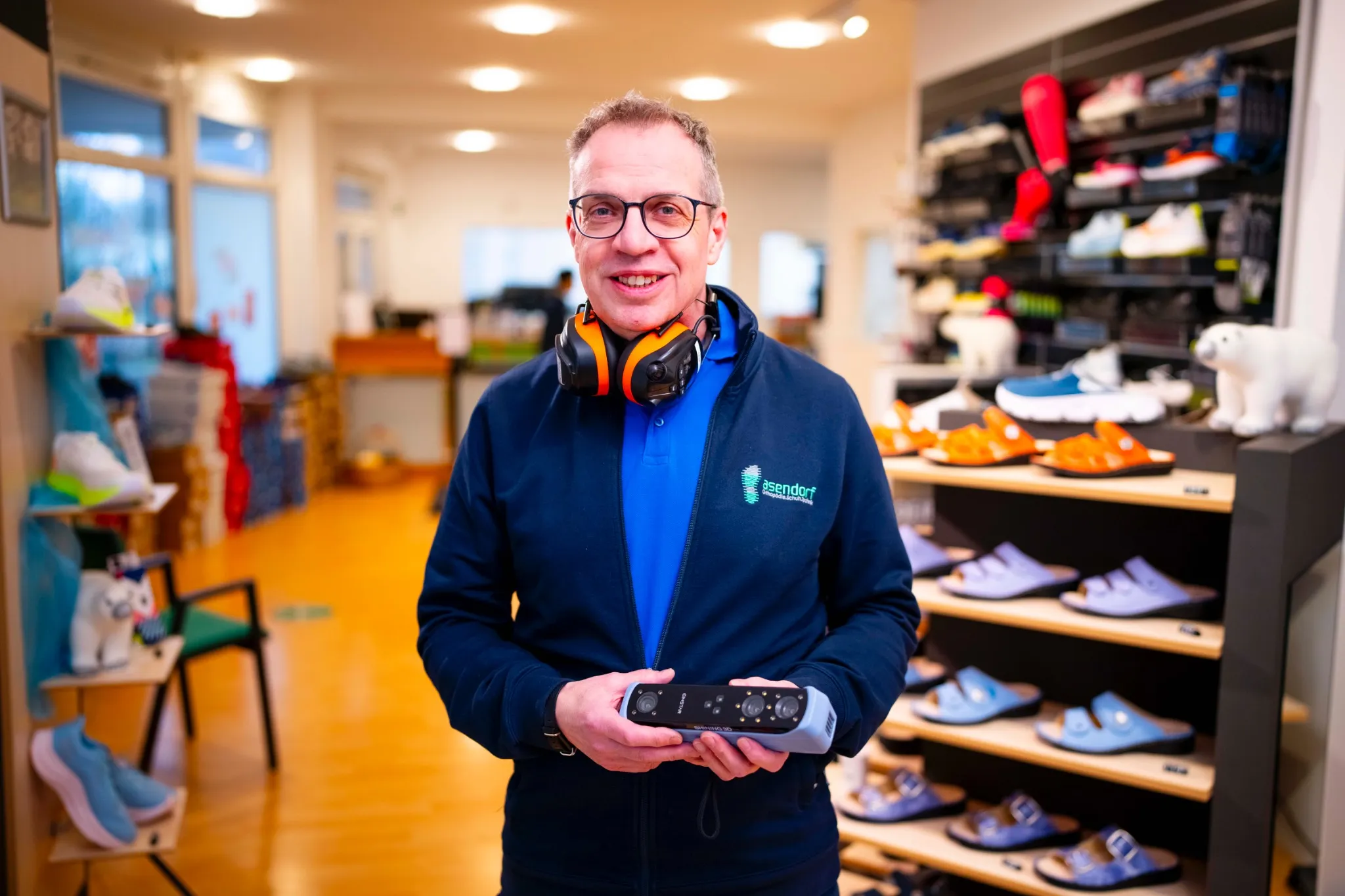 Jens Asendorf mit Kopfhörern um den Hals steht in einem Schuhgeschäft und hält ein Handheld-Gerät in der Hand. Im Hintergrund sind Regale mit Schuhen und ein ausgestopfter Eisbär zu sehen.