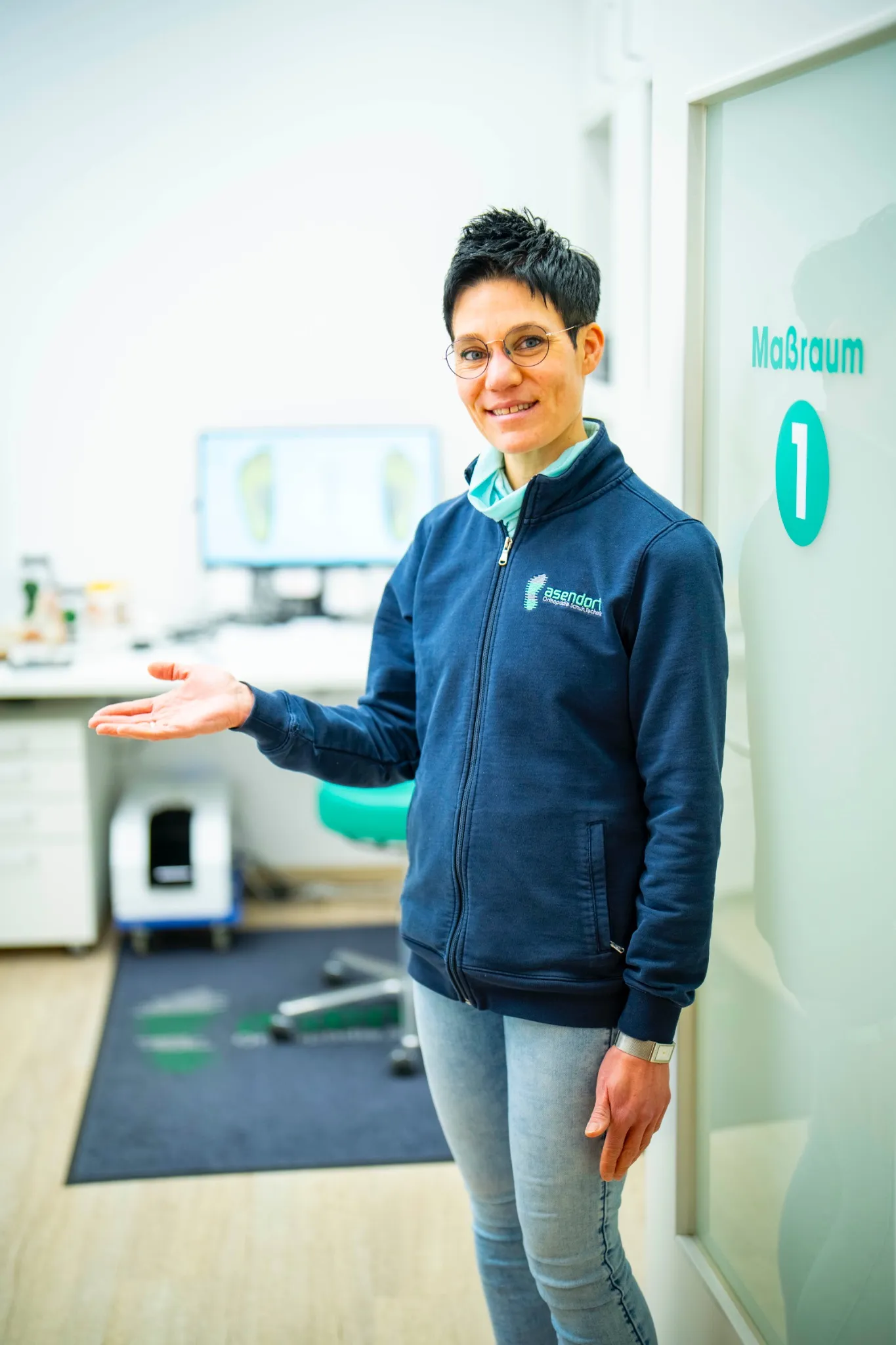 Eine Frau in einer blauen Jacke steht in einem Innenraum, lächelt und deutet auf einen Raum mit der Aufschrift "Maßraum 1", in dem im Hintergrund medizinische oder Bürogeräte zu sehen sind.