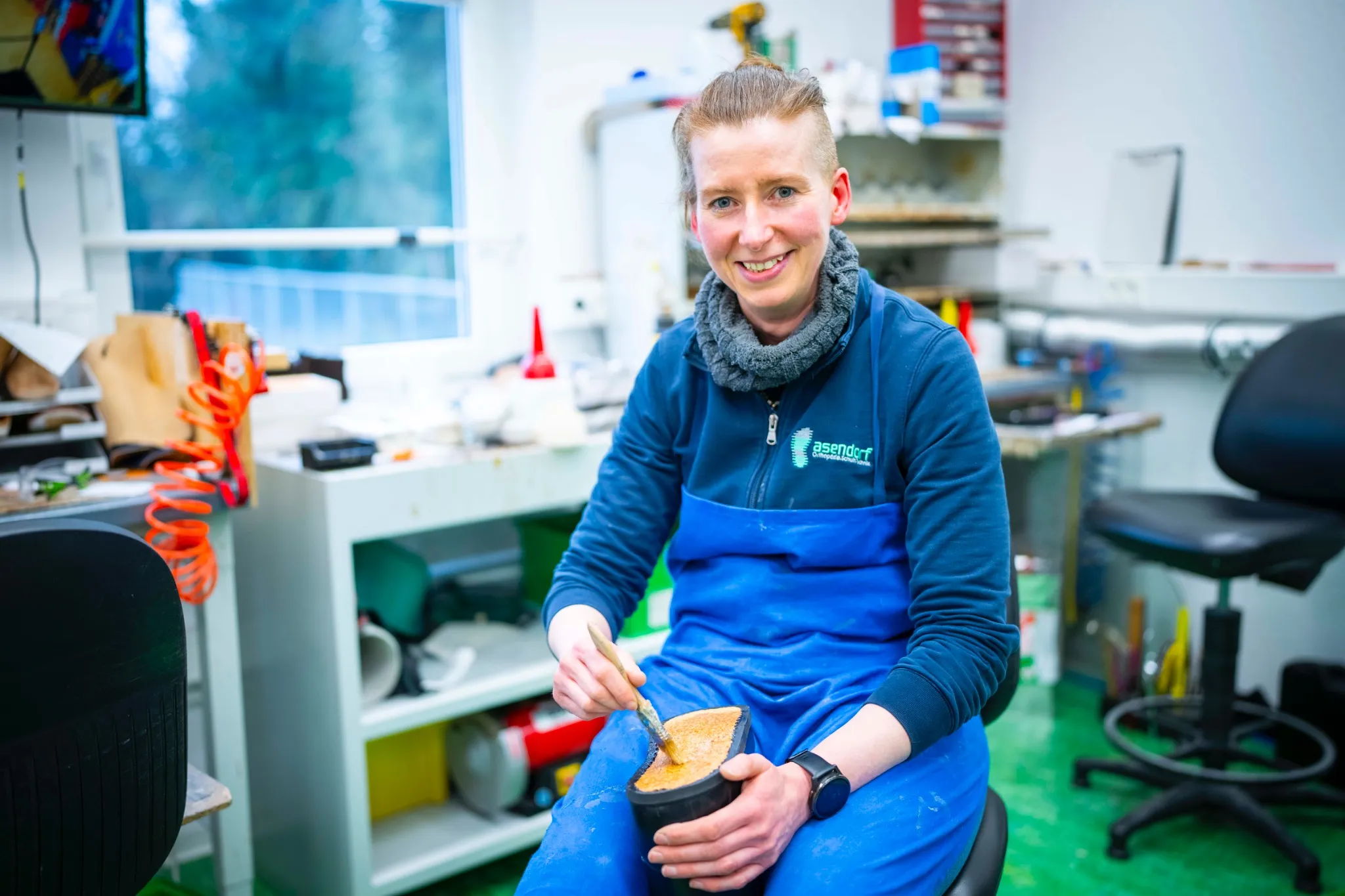 Eine Frau in blauer Schürze sitzt in einer Werkstatt, hält ein Werkzeug in der Hand und formt ein Stück Material, wobei im Hintergrund verschiedene Werkzeuge und Geräte zu sehen sind.