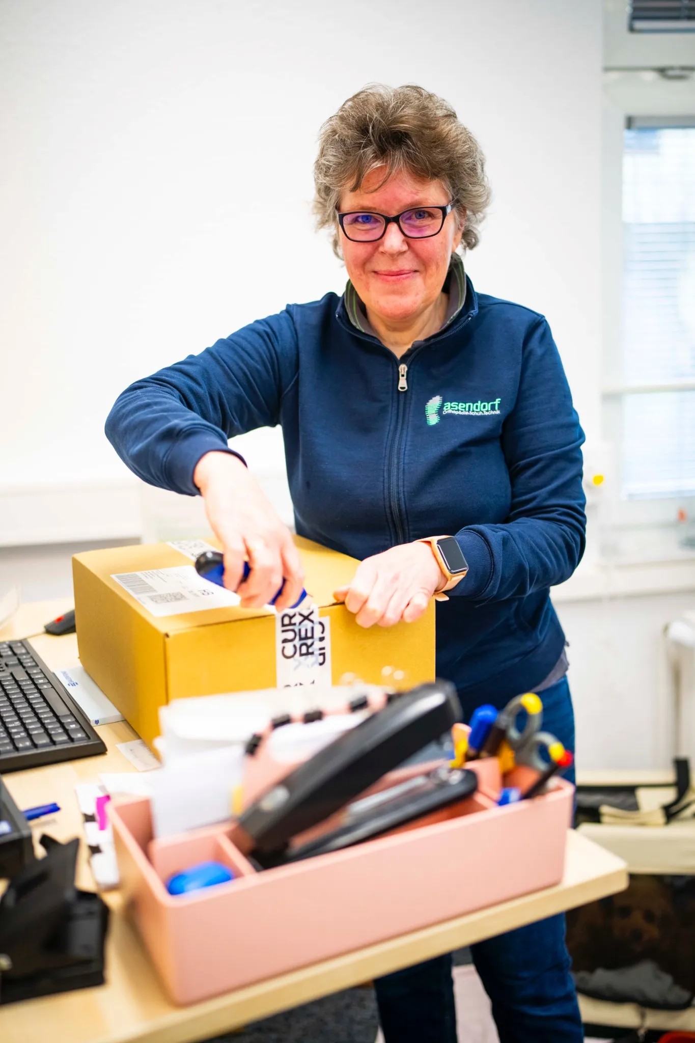 Eine Frau in einer blauen Jacke öffnet einen gelben Karton mit einem Kartonschneider an einem Bürotisch mit verschiedenen Materialien.