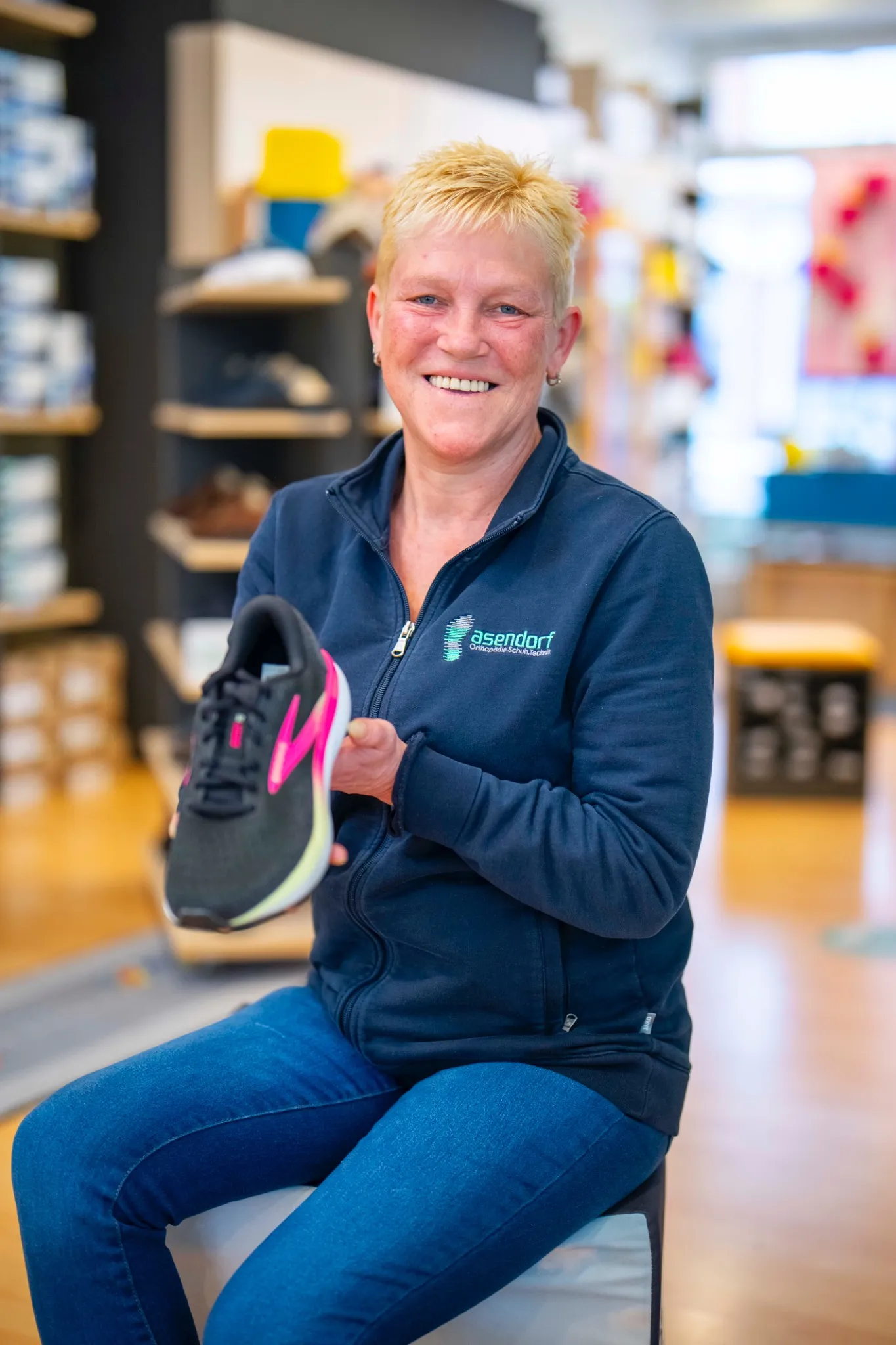 Eine Frau mit kurzen blonden Haaren, die eine marineblaue Jacke und Jeans trägt, sitzt in dem Schuhfachgeschäft Asendorf und hält einen schwarzen Sportschuh mit rosa Akzenten hoch.