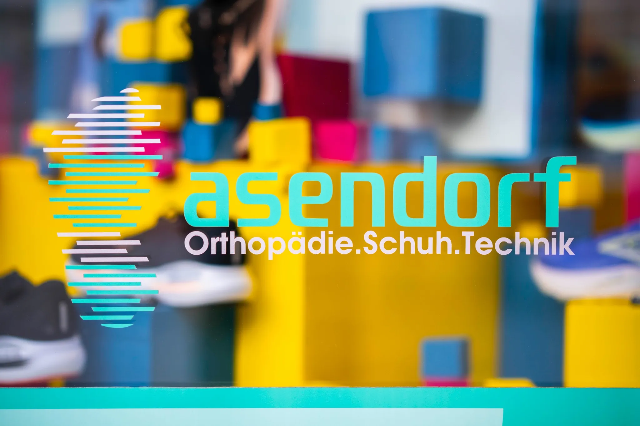 Das Bild zeigt ein Schild mit der Aufschrift "asendorf Orthopädie.Schuh.Technik" auf einem Glasfenster mit verschwommenen bunten Blöcken und Schuhen im Hintergrund.