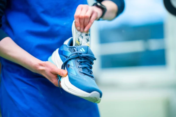 Eine Person in einer blauen Schürze legt eine gemusterte Einlegesohle in einen blauen Sportschuh ein, wobei sie den Schuh mit einer Hand und die Einlegesohle mit der anderen hält.