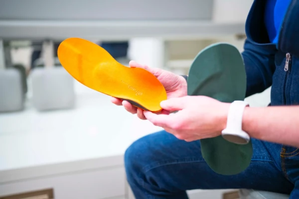 Eine Person, die eine Smartwatch trägt, hält zwei verschiedenfarbige orthopädische Einlagen, eine orange und eine grün, in einer hellen Innenumgebung.