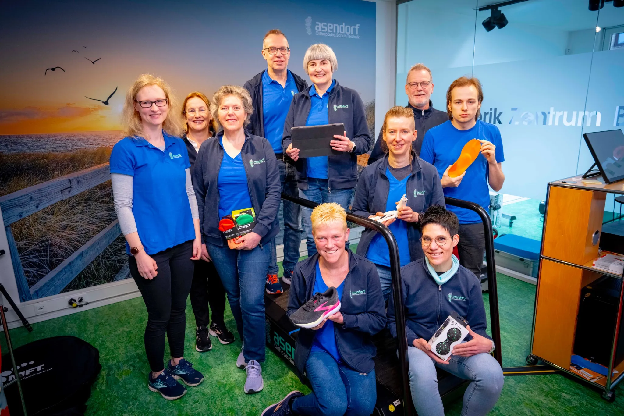 Eine Gruppe von zehn Personen in blauen und schwarzen Uniformen posiert in einem Innenraum mit Schuheinlagen und Ausrüstung um ein Laufband herum.